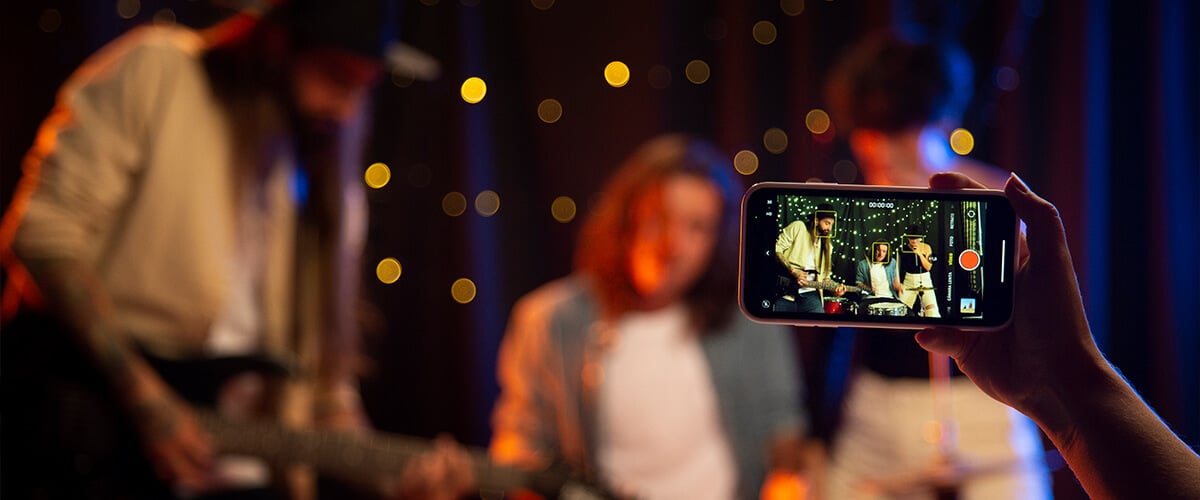 Hand holding a smartphone recording a live concert performance with band members in the background.