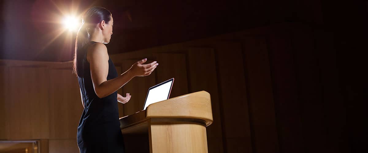 A female business executive giving a speech at a podium with a spotlight shining behind her, presenting to an audience in a professional setting.
