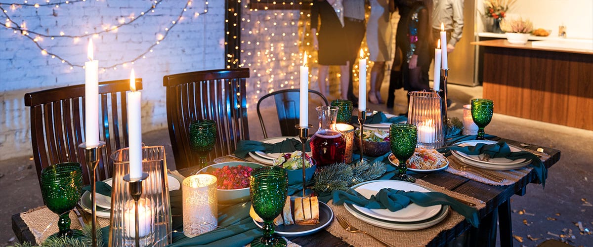 Festive New Year party table setting with candles, green glasses, and a variety of dishes, surrounded by warm lighting and family members in the background.