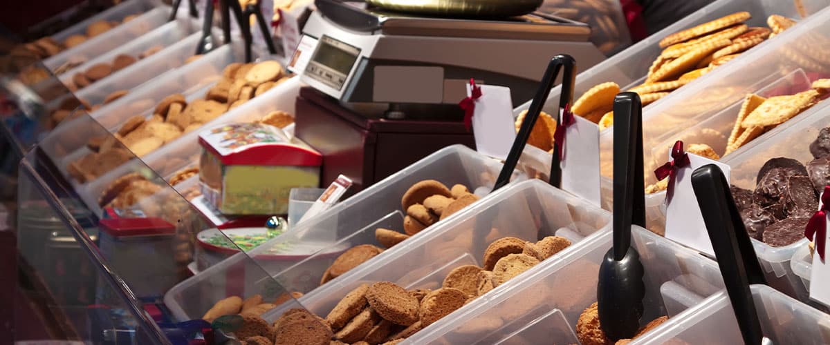 Assorted pastries and cookies displayed in plastic containers on a market counter with tongs and price tags.