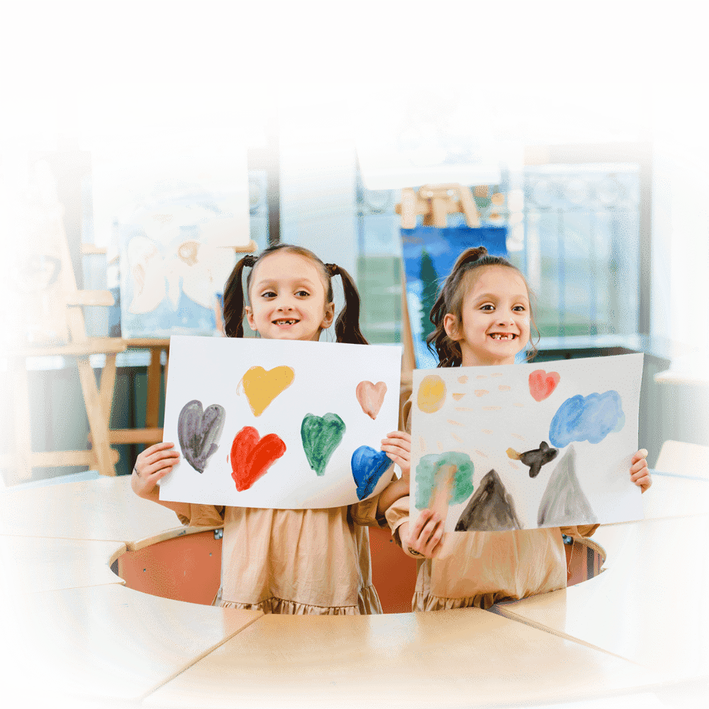 Two young girls with pigtails proudly holding up their colorful artwork featuring hearts, nature, and abstract elements in a classroom setting.